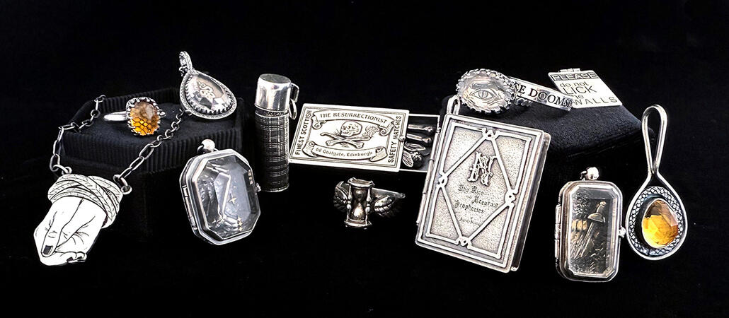 A group of sterling silver jewelry against a black background, from left to right: a necklace with a clasped pair of hands bound by rope at the wrists; a ring with a cushion-cut citrine set over a snakeskin texture; a faceted teardrop crystal quartz set ov